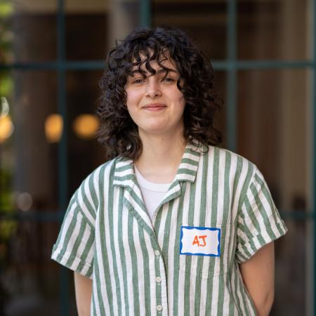 AJ Damiana wears a striped button-up with a smile, her dark, curly hair falling to her shoulders.